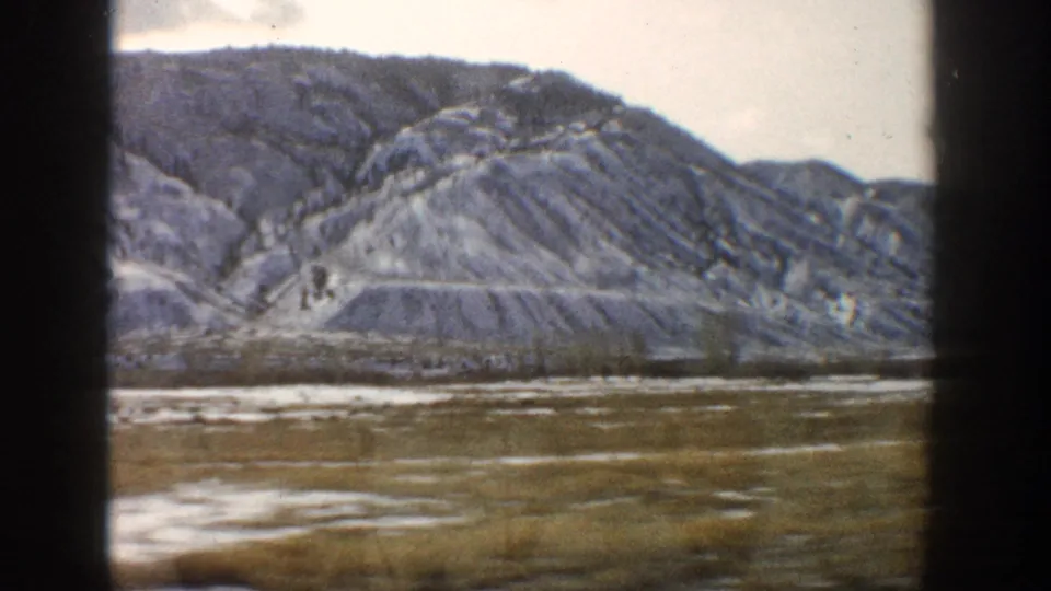 1961 Aspen, Colorado home-movie frame with a broad valley foreground and rugged snow-streaked mountain slope seen from a moving vehicle.