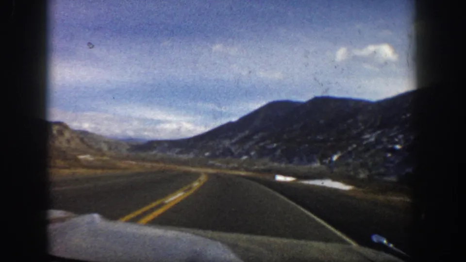 1961 Aspen, Colorado road-travel view from a car, with a curved paved road, valley floor, rocky slopes, and bright snow patches.