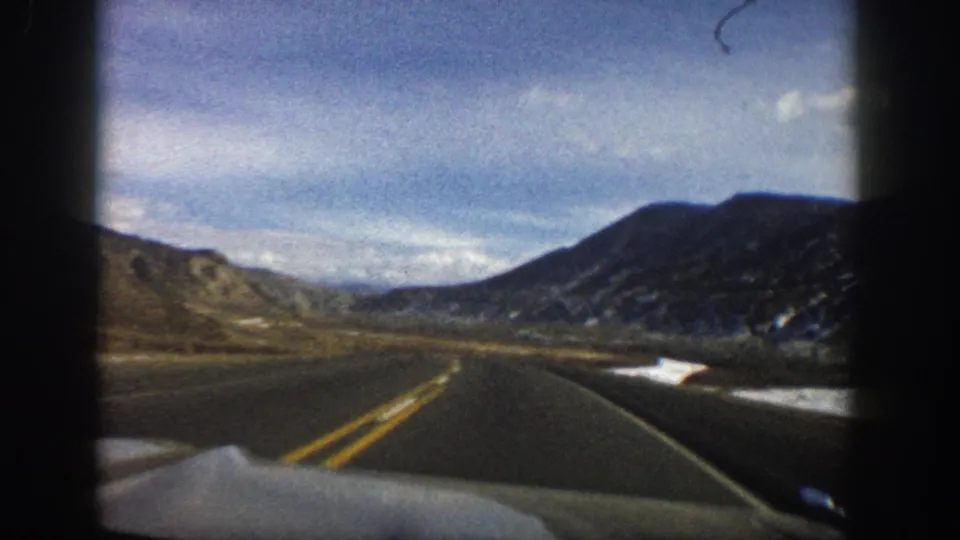 1961 Aspen, Colorado 8mm frame with a forward car view of a two-lane road, bare slopes, distant valley, snow, and clouded sky.