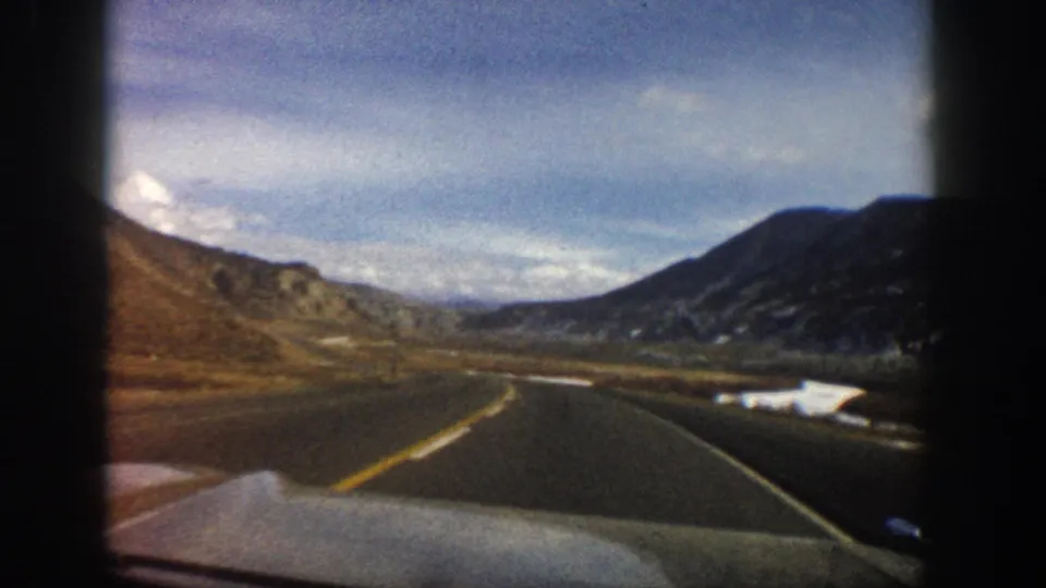 1961 Aspen, Colorado home-movie still showing a road curving through a bare mountain valley under a blue, lightly clouded sky.