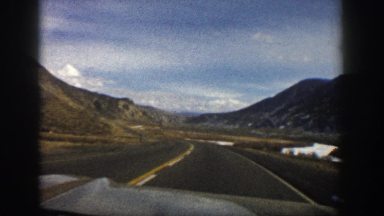 1961 Aspen, Colorado road-trip view from inside a moving car, showing a paved highway between arid hills and distant mountains.