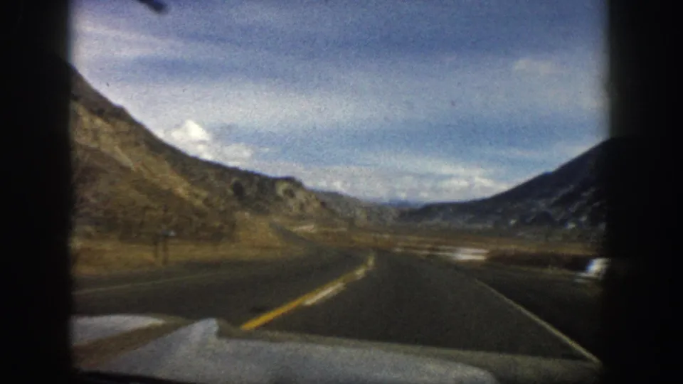 Mid-century 8mm view near Aspen, Colorado, with a road bending through hills under a blue sky and scattered clouds.