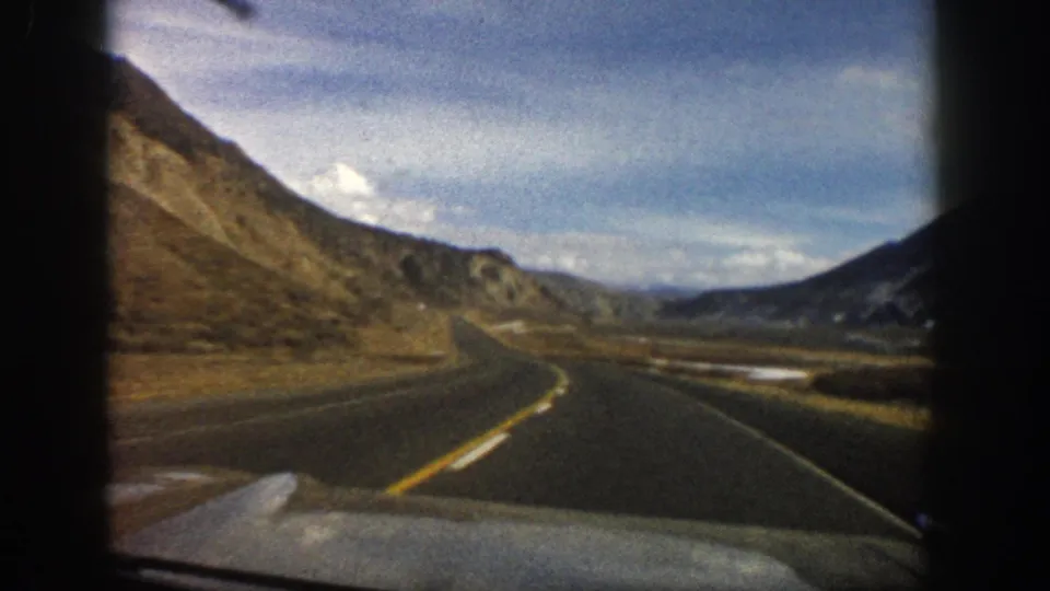 Aspen-area 1961 home-movie frame showing a paved mountain road, windshield edge, dry slopes, and distant ridgelines.