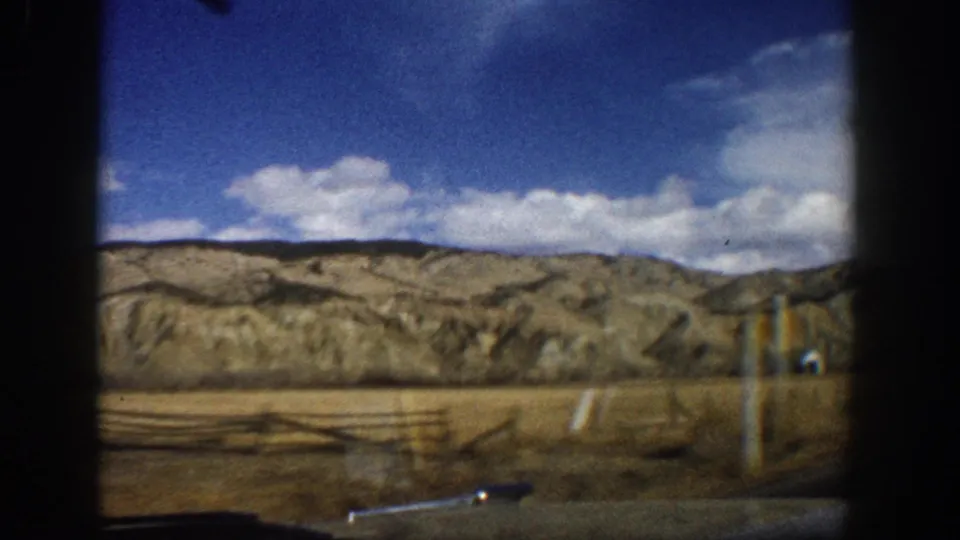 1961 8mm road-view frame with fence posts, dry grassland, distant hills and a small roadside structure far back.