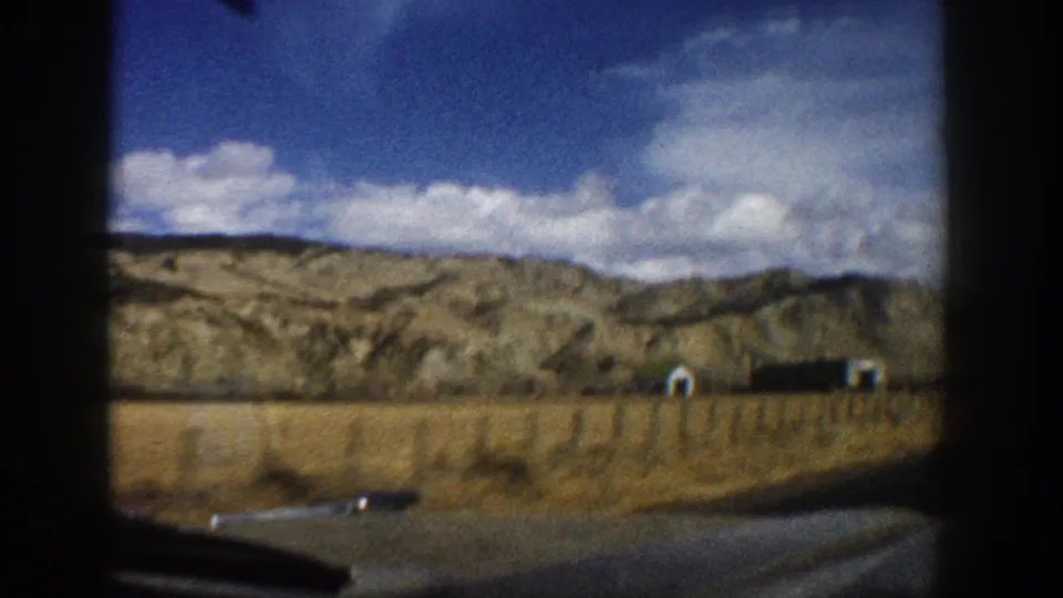 Aspen-area home-movie frame showing dry roadside land, distant small buildings, hills and a cloudy blue sky from a moving car.
