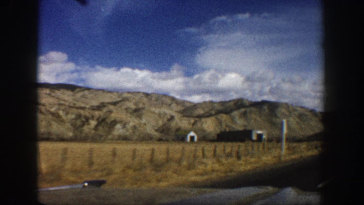 1961 Aspen, Colorado home-movie frame showing dry terrain, small roadside structures, and rolling brown mountains from a moving car.