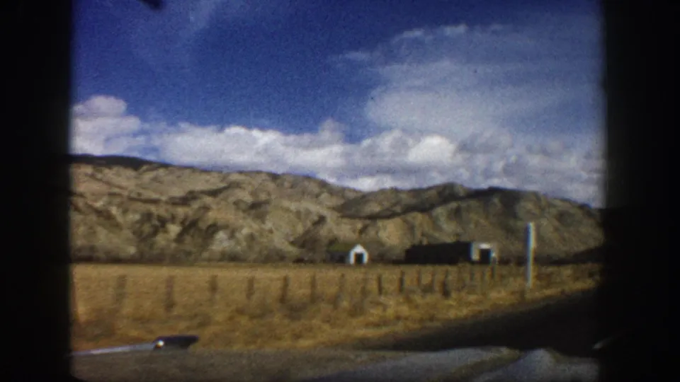 1961 Aspen, Colorado home-movie frame showing dry terrain, small roadside structures, and rolling brown mountains from a moving car.