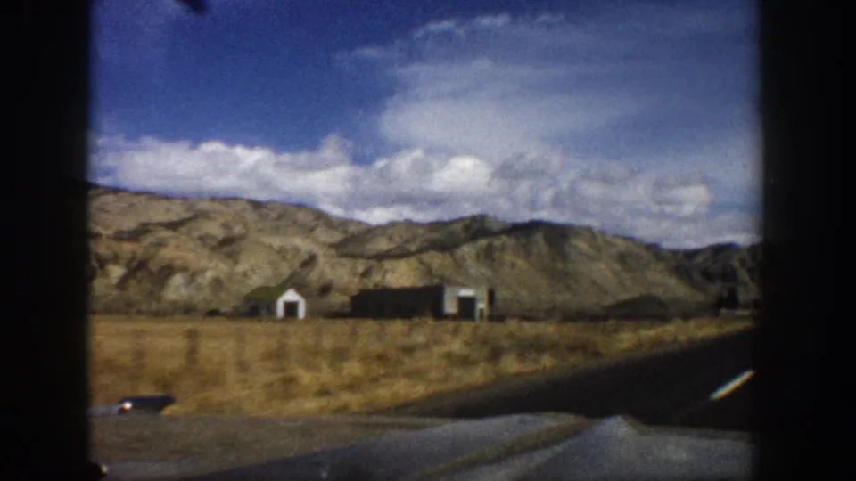 Passenger-view frame from 1961 Aspen, Colorado, with roadside structures, dry grassland, mountains, and a paved road edge.