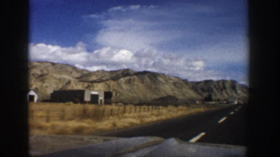 1961 Aspen, Colorado road-travel frame showing a highway, arid field, small roadside buildings, and mountain ridges under clouds.