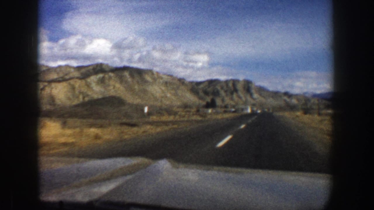 1961 Aspen-area 8mm road view from a moving car, showing asphalt lanes, a car hood, dry hills, and a bright cloudy sky.