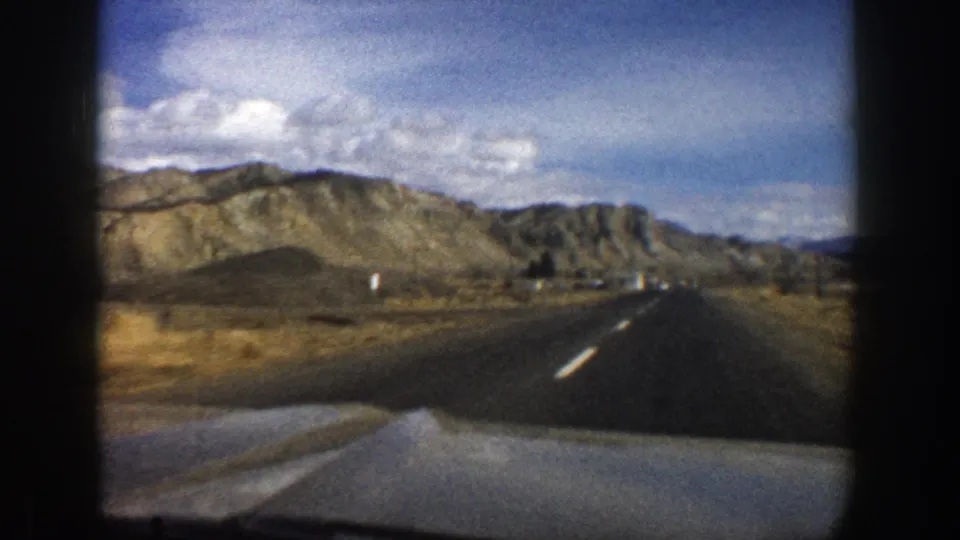 1961 Aspen-area 8mm road view from a moving car, showing asphalt lanes, a car hood, dry hills, and a bright cloudy sky.