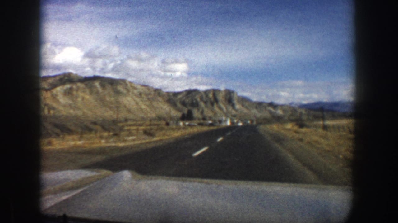 Road curves through a dry mountain landscape with hills and distant structures.