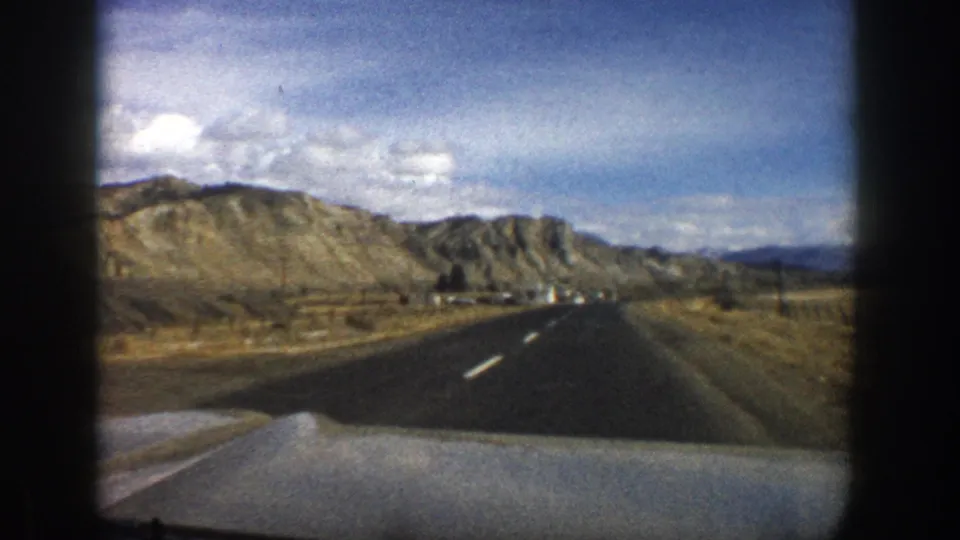 Moving-car view near Aspen, Colorado in 1961, with a paved road, golden roadside terrain, rocky hills, and small distant structures.