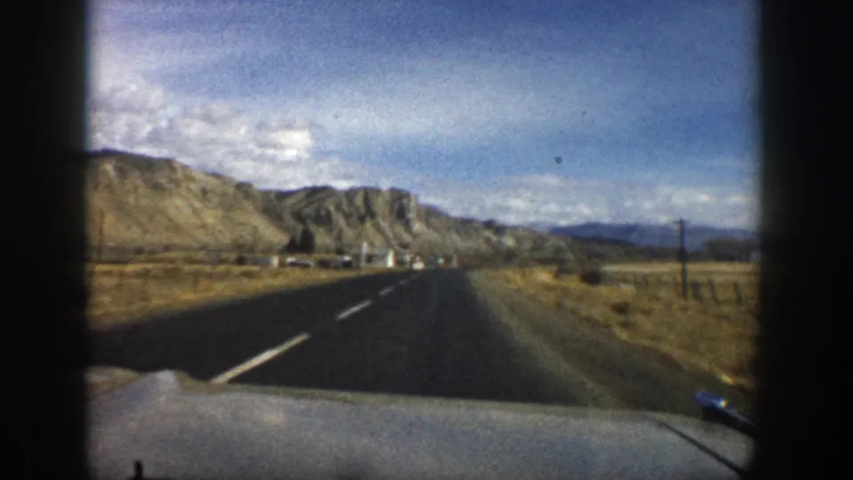8mm home-movie frame from a moving vehicle near Aspen in 1961, showing a paved road, roadside poles, dry grass, and mountain ridges.