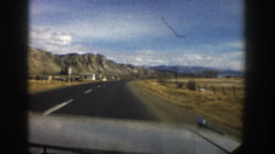 1961 Aspen-area driving footage shows a two-lane road, car hood, roadside fencing or posts, dry grass, and layered mountain ridges.
