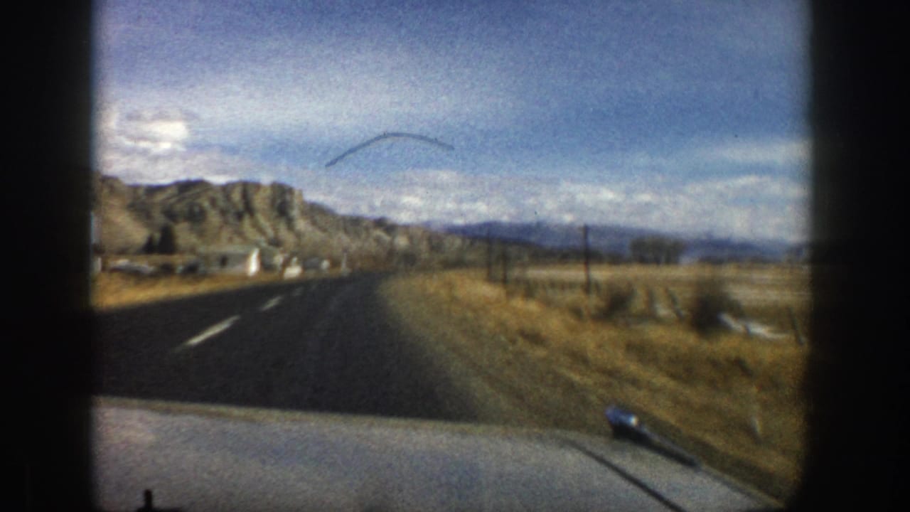 1961 Aspen-area 8mm frame from a moving car on a rural road toward rocky Colorado hills and open fields.