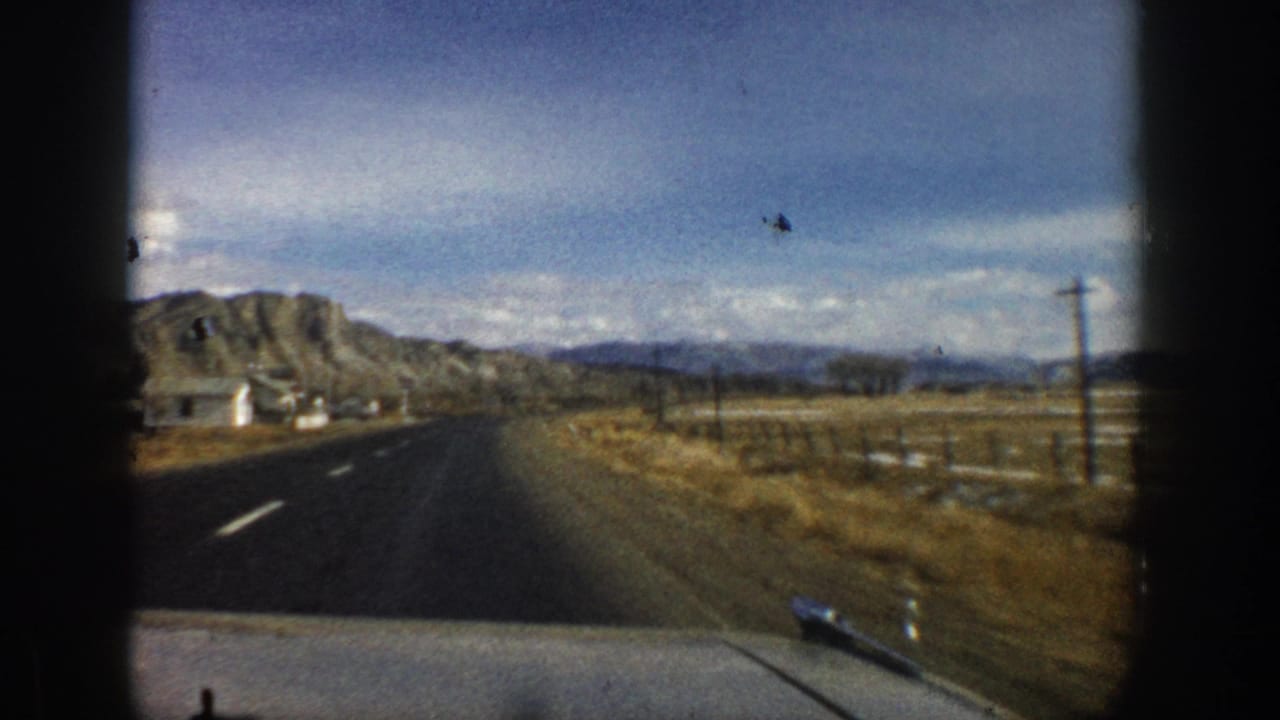 1961 Aspen-area road scene showing a small roadside structure, open fields, poles, and mountain ridges from a moving vehicle.