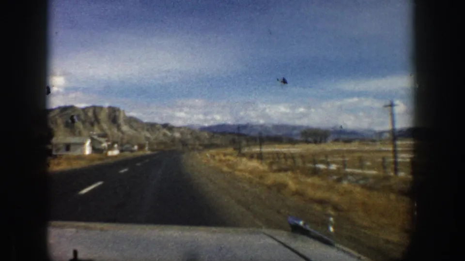 1961 Aspen-area road scene showing a small roadside structure, open fields, poles, and mountain ridges from a moving vehicle.