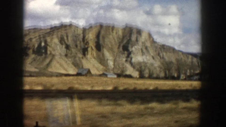 1961 Aspen-area 8mm view of a rocky mountainside rising beyond dry grassland and small rural buildings.