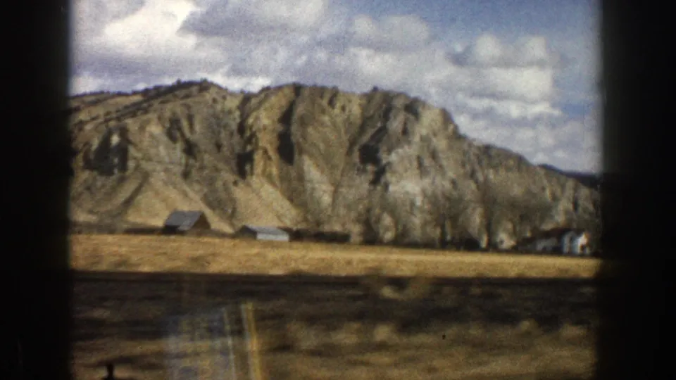 1961 Aspen-area countryside frame with a rugged hillside, open field, low buildings, and bare trees under broken clouds.