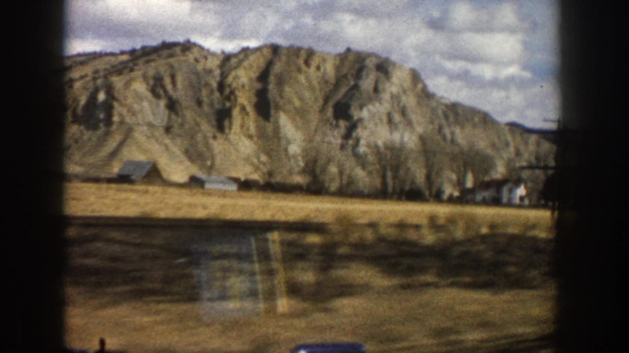 1961 Aspen-area 8mm frame showing rocky cliffs, dry fields, scattered buildings, and a cloudy mountain sky from a moving viewpoint.