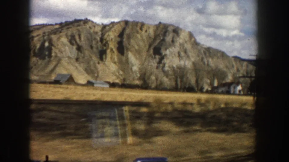1961 Aspen-area 8mm frame showing rocky cliffs, dry fields, scattered buildings, and a cloudy mountain sky from a moving viewpoint.