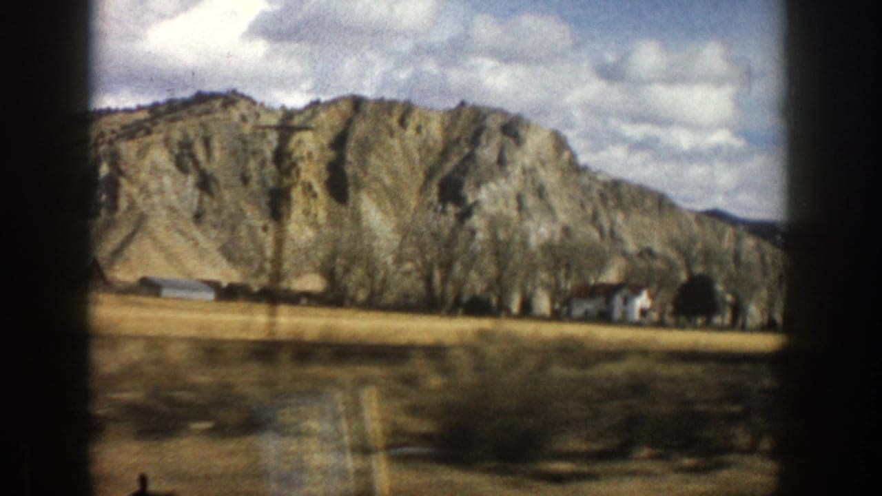 Mountain cliff, open field, small structures, and cloudy sky seen through dark frame edges.