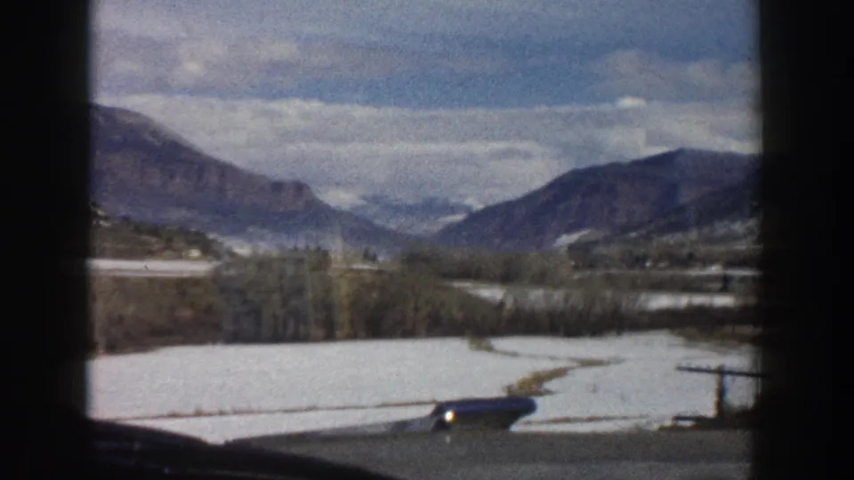 1961 Colorado mountain travel frame showing a snowy valley, bare trees, distant ridges, and road foreground from a moving vehicle.