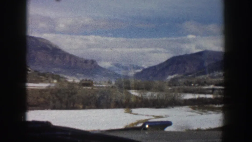 Aspen-area 8mm frame showing snow patches, a winding road or track, bare trees, and distant mountain ridges under cloud cover.