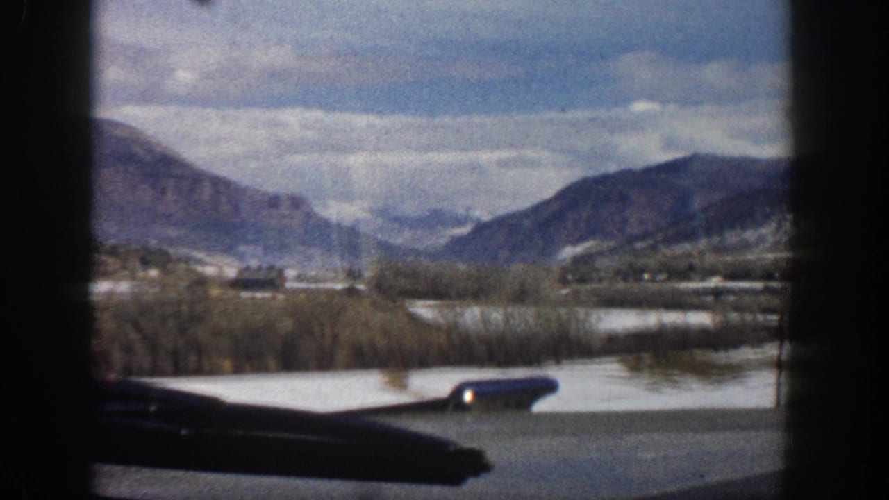 1961 Aspen-area mountain valley viewed from a moving car window, with snow patches, bare trees, distant ridges, and a side mirror visible.