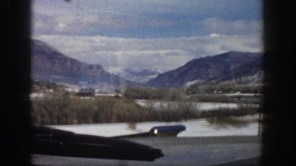1961 Aspen-area mountain valley viewed from a moving car window, with snow patches, bare trees, distant ridges, and a side mirror visible.