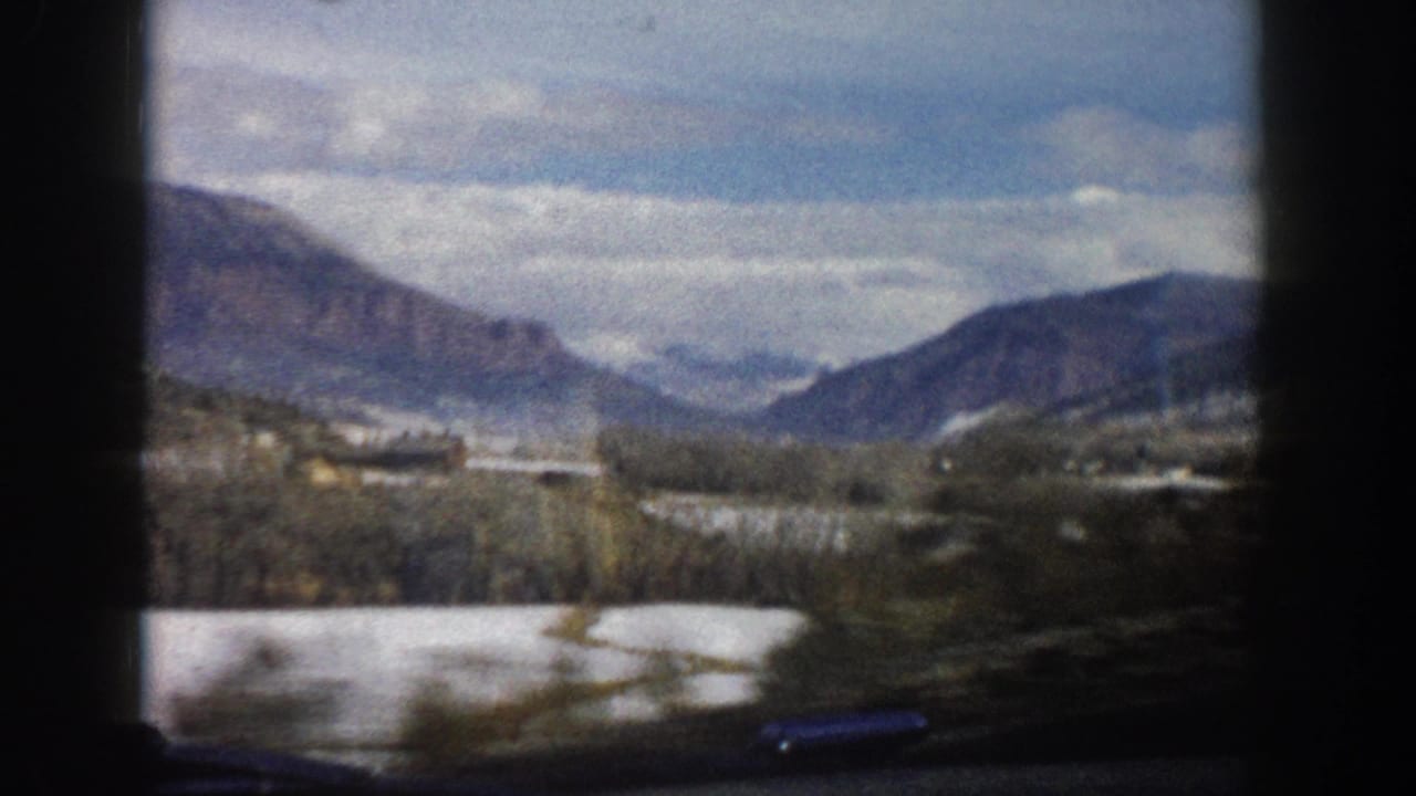 Aspen, Colorado valley landscape from a moving car in 1961, with snow-covered ground, bare trees, low buildings, and mountains beyond.