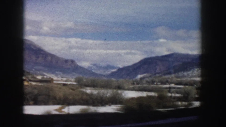 Soft 8mm frame of a Colorado mountain valley from a moving car, showing snow-dusted fields, bare trees, distant peaks, and interior framing.