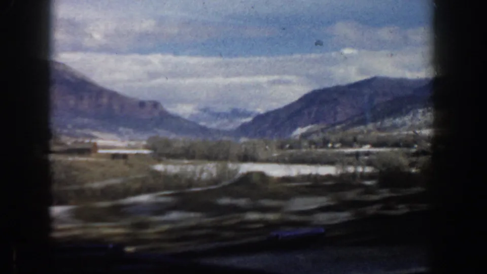 Late clip frame from a 1961 Aspen car ride, showing a curving road, snow patches, valley trees, and layered mountain ridges through the window.