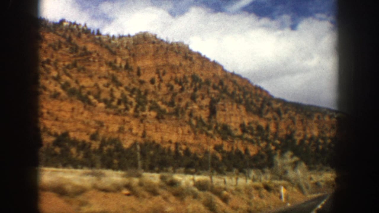 1961 Aspen-area mountain road view with reddish slopes, scattered evergreens, clouds, and car-window framing.