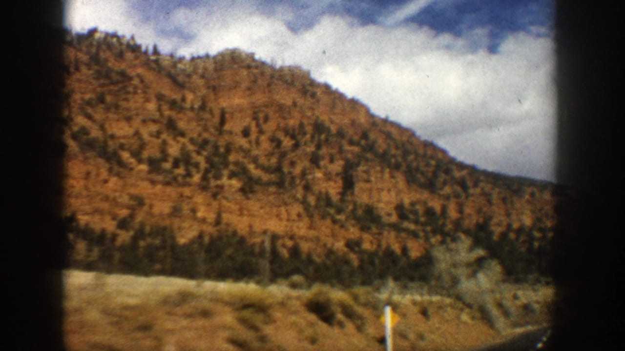 Aspen, Colorado road footage from 1961 showing a reddish hillside, evergreens, cloudy sky, and yellow roadside sign.