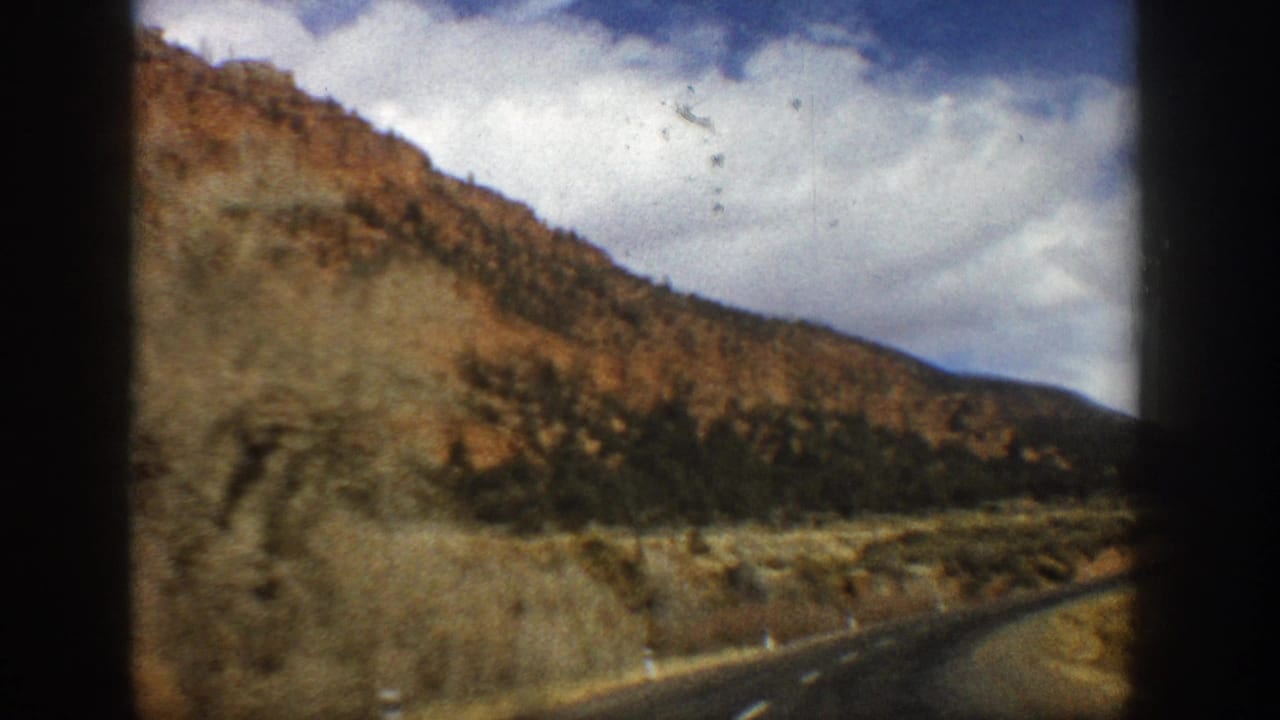 1961 Aspen, Colorado home-movie frame showing a paved road curving past reddish mountain slopes, scrub, and clouds.
