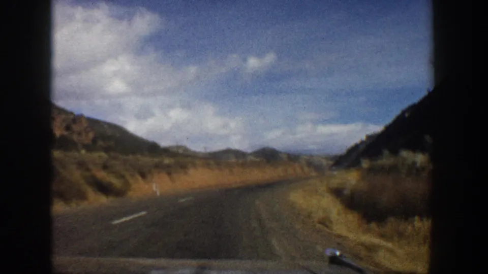 End frame of a 1961 Aspen, Colorado mountain drive, with an empty road curving between hills and dry roadside vegetation.
