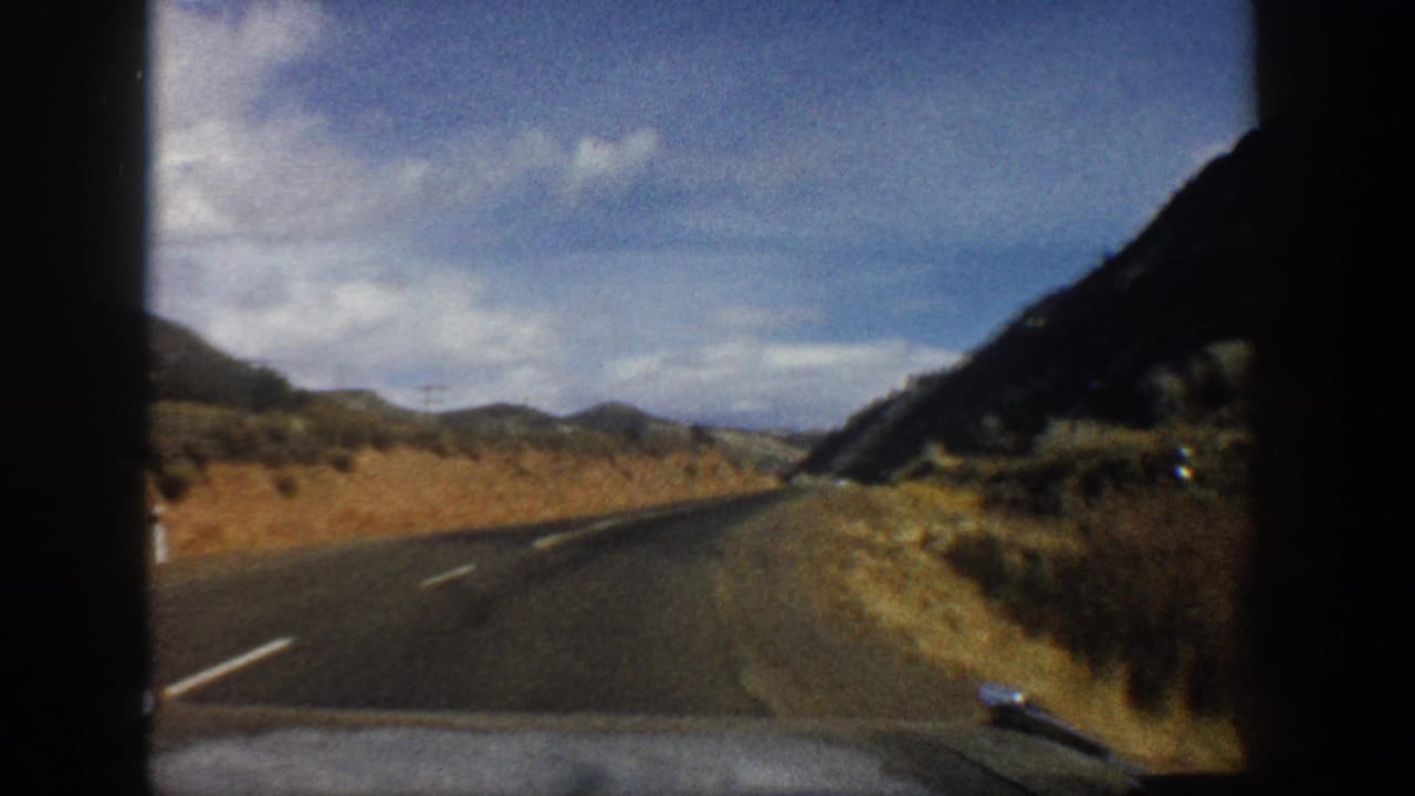1961 Aspen-area 8mm view from a car approaching a valley road bordered by dry slopes and mountain terrain.