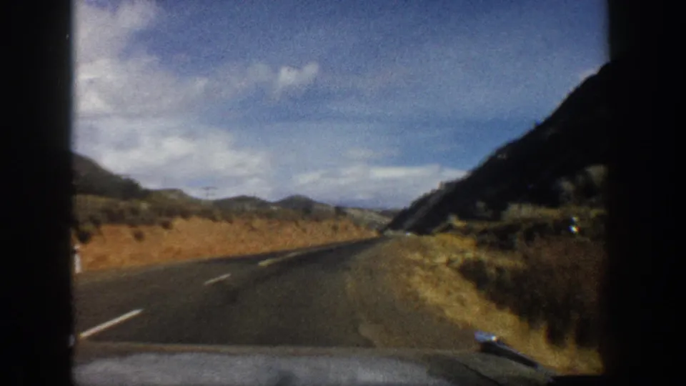 1961 Aspen-area 8mm view from a car approaching a valley road bordered by dry slopes and mountain terrain.
