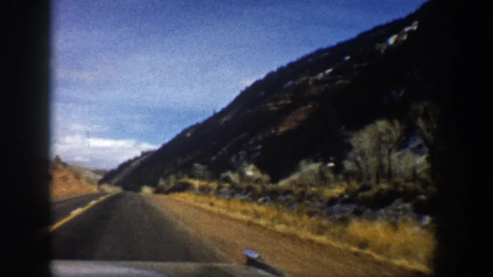 Aspen-area 1961 home-movie frame showing a car traveling along a two-lane road below a rocky mountain slope.