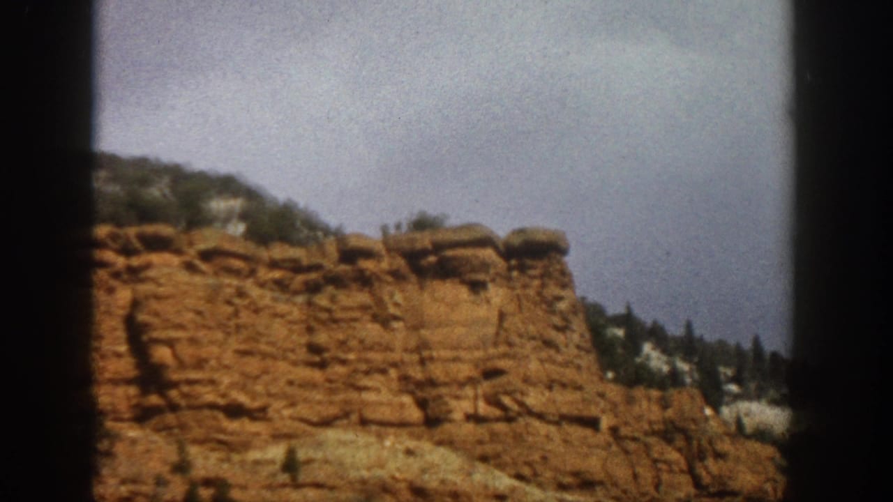 1961 Aspen, Colorado home-movie frame showing a stratified red rock cliff, treed ridge, and pale overcast sky through dark borders.