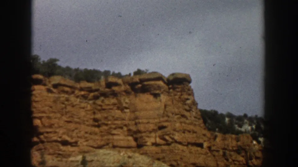 Aspen, Colorado 1961 home-movie frame of a weathered red cliff face, ridge trees, cloudy sky, and soft 8mm texture.