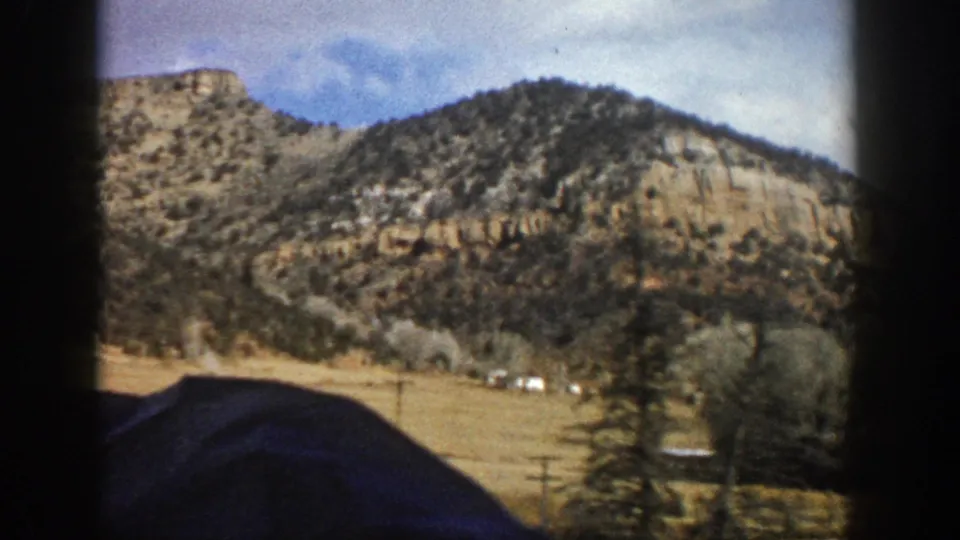 1961 Aspen, Colorado home-movie frame with a mountain ridge, dry field, scattered trees, and a dark foreground car-window shape.