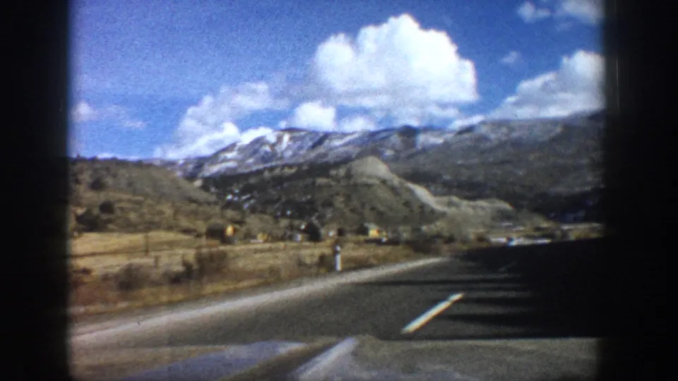 1961 Aspen, Colorado road view from a moving car, with valley land, distant snow-dusted mountains, blue sky, and clouds.