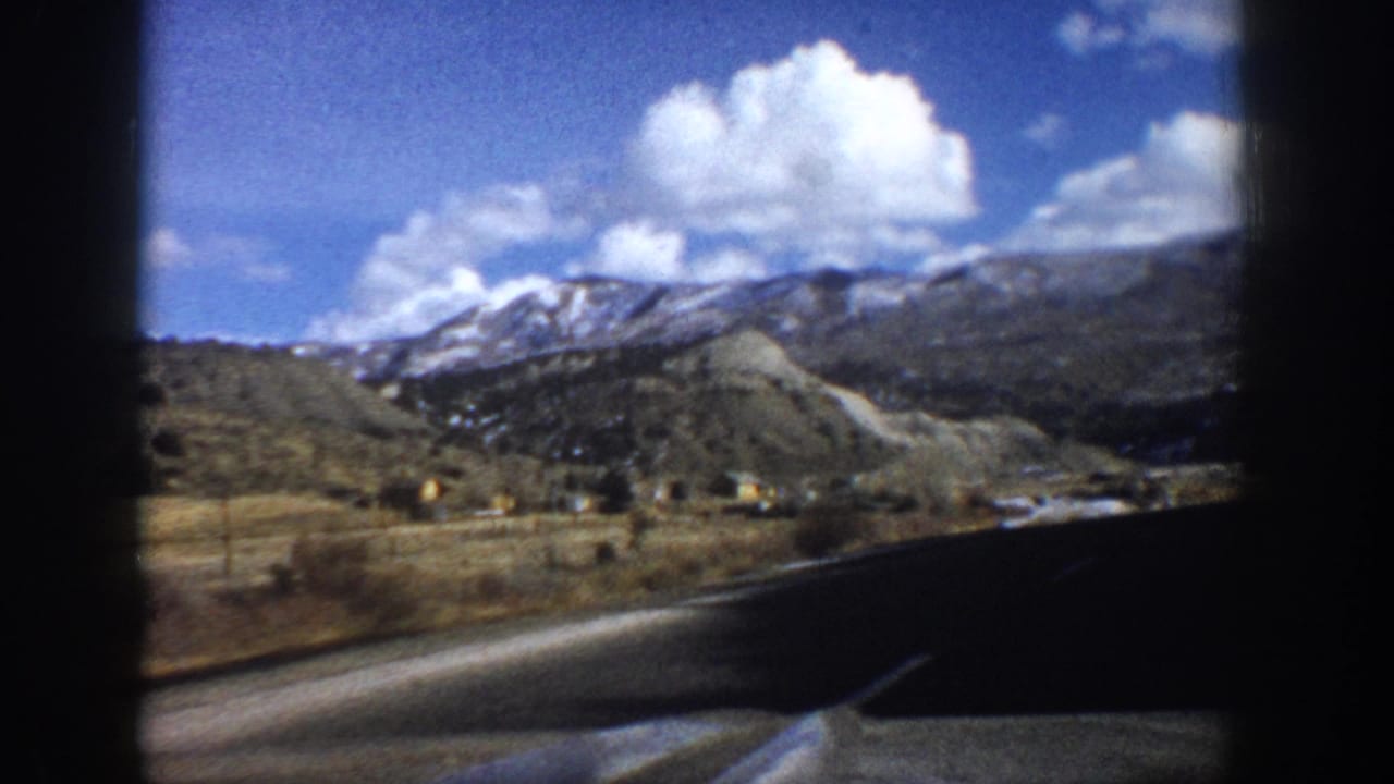 1961 Aspen-area mountain road seen from a moving car, with snowy ridges, open valley ground, clouds, and dark 8mm frame borders.