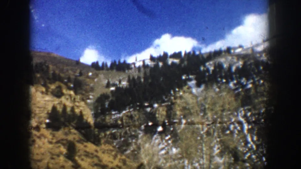 Moving-car view of a Colorado mountain hillside near Aspen, with dark evergreens, exposed winter brush, snow traces, and 8mm film grain.