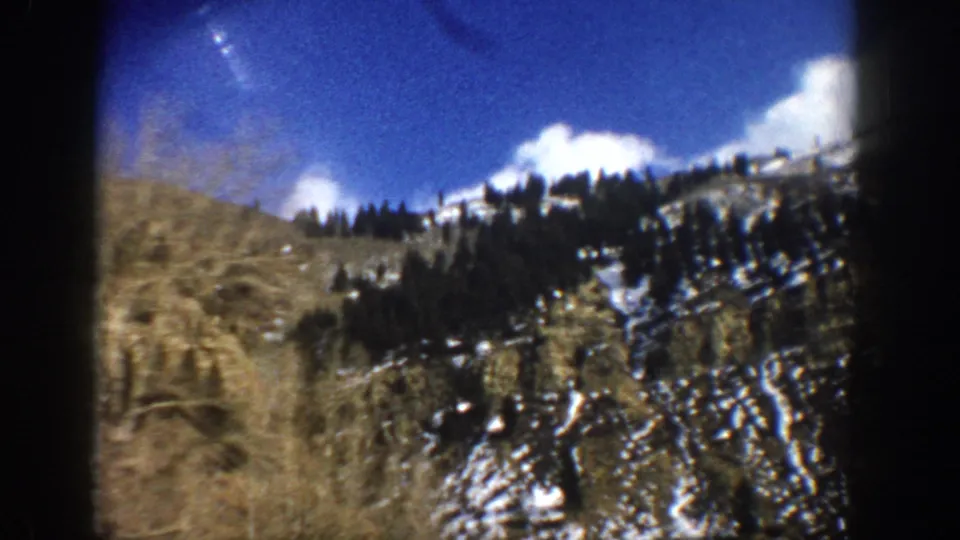 Final 1961 Aspen-area frame showing a snow-streaked evergreen hillside, bare foreground brush, blue sky, and dark projected-film borders.