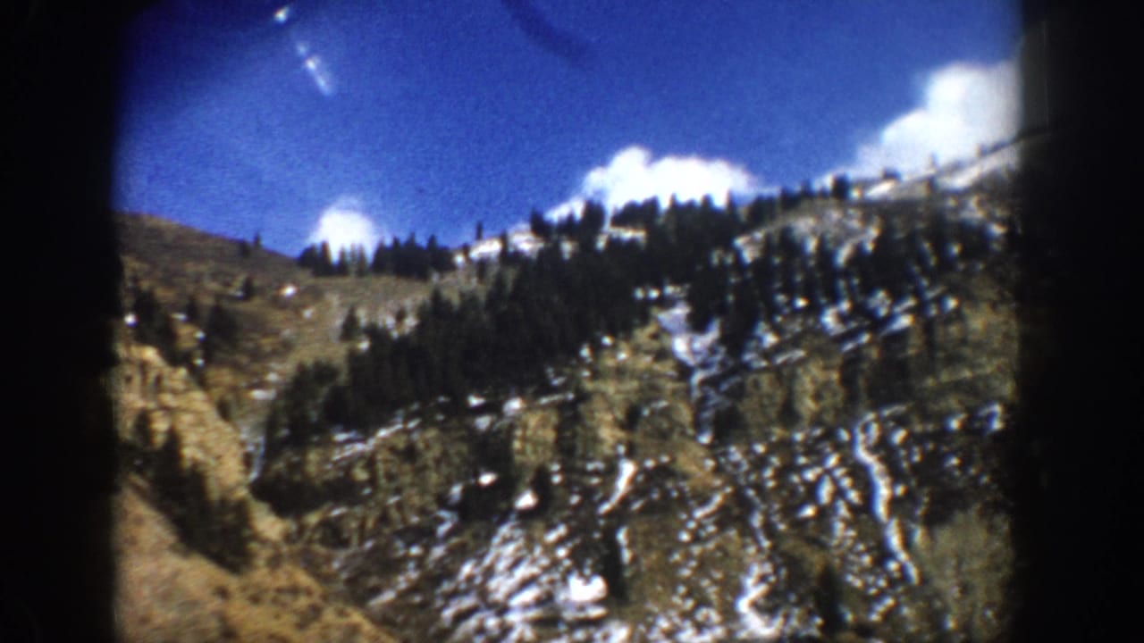 1961 Aspen-area 8mm frame showing a snow-dusted rocky slope, evergreen trees, blue sky, and dark film-edge vignetting.
