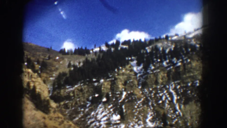 1961 Aspen-area 8mm frame showing a snow-dusted rocky slope, evergreen trees, blue sky, and dark film-edge vignetting.